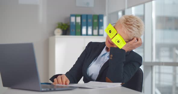 Tired Mature Woman Sleeping at Workplace with Stickers on Her Eyeglasses