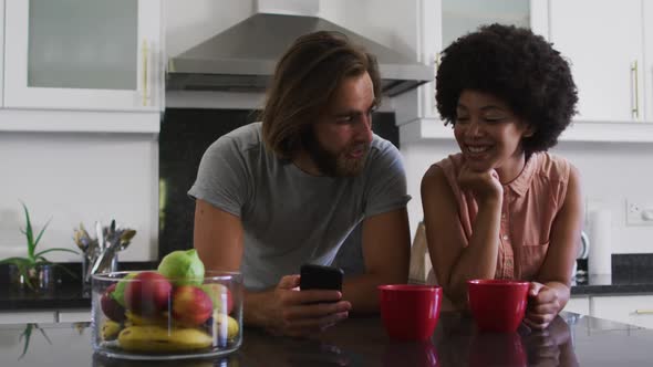 Mixed race couple holding coffee cup using smartphone in the kitchen at home