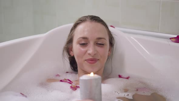 Portrait of Relaxed Smiling Beautiful Woman Lying in Water in Bathtub Blowing Out Candle in Slow