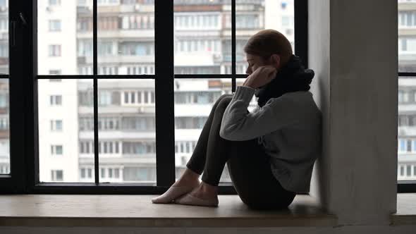 Young woman is sad sitting on the windowsill of city apartment