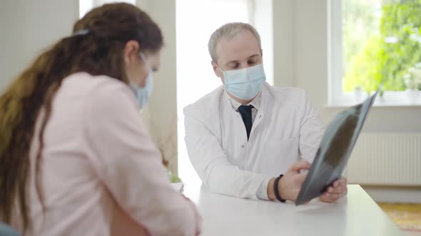 Caucasian Doctor in Face Mask Explaining Covid-19 Complications on Lungs X-ray To Patient. Portrait