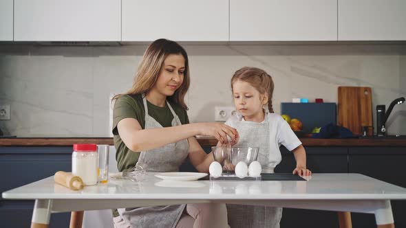 Mother with Little Daughter Cracks Egg Into Bowl at Table