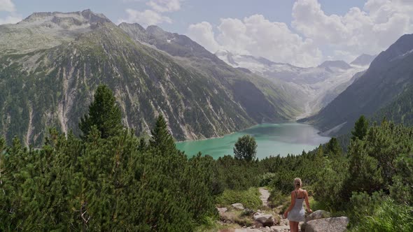 A caucasian girl in a white dress walks down a path towards a blue lake and a beautiful mountain sce