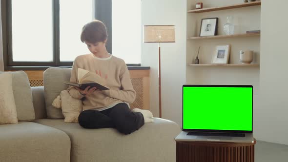 A Laptop with a Green Screen Stands on a Table Against of a Woman Reading a Book