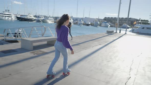 Lone Darkhaired Girl Roller Skates in Sunlight By Boats in Harbor