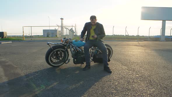 Young Man with Smart Phone in Hands on Custom Bobber Motorcycle on Street at Sunset Slow Motion