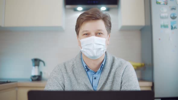 Portrait of a Young Attractive Man in a Medical Mask at Home at a Computer