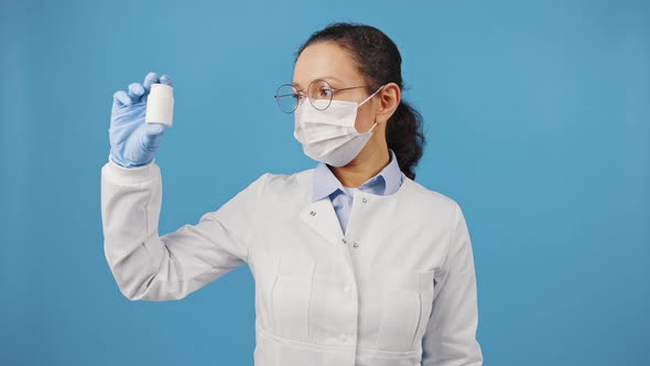 Professional Woman Doctor Wearing Protective Mask Showing Jar with Pills at Camera Blue Studio