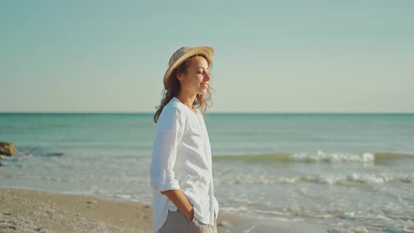 Attractive Smiling Woman Traveler in White Shirt and Straw Hat Walking Along Sea Coastline