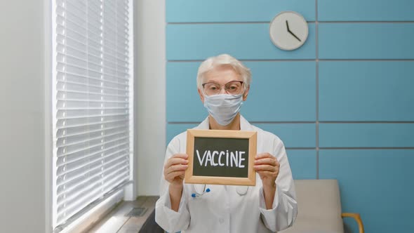 Grey haired lady doctor with protective mask holds blackboard with word Vaccine