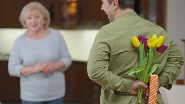 Back View Man Hiding Bouquet of Flowers and Gift Behind Back Congratulating Woman on Birthday or