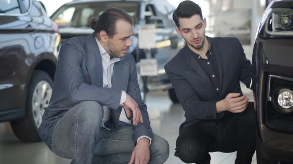 Young Caucasian Trader Sitting on Hunkers with Male Client and Showing Car Wheel