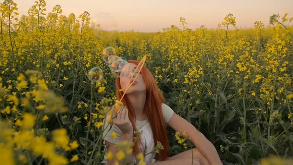 Young Redhair Woman Blowing Bubbles at the Camera Outdoors in Summer Meadow