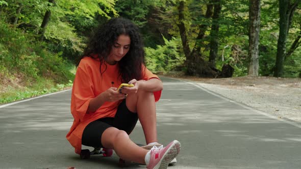 Sporty Girl Using Mobile Sitting on Skateboard in the Park