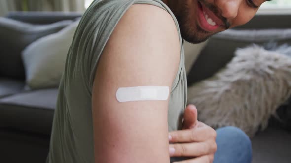 Smiling young man looking at bandage on arm while sitting at home after vaccination
