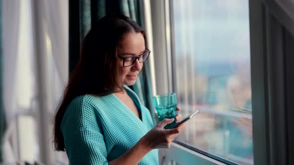 In the Morning a Beautiful Woman is Standing at the Window with Glasses Watching the News on Her