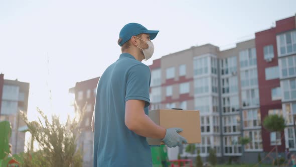 The Male Deliveryman in a Cap and a Protective Mask and Gloves Goes with a Box in His Hands and