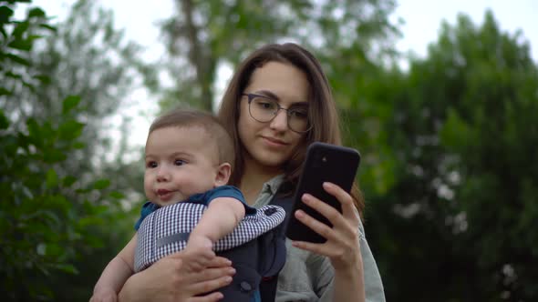 A Young Woman with a Child Stands with a Phone in Her Hands in the Park