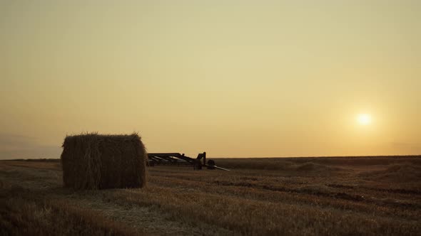 Hay Bale on Wheat Field with Harvesting Equipment at Golden Sunset