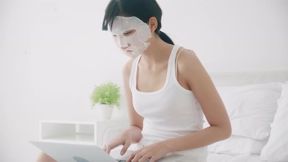 Beautiful young asian woman with sheet facial mask sitting working on laptop computer on bed.