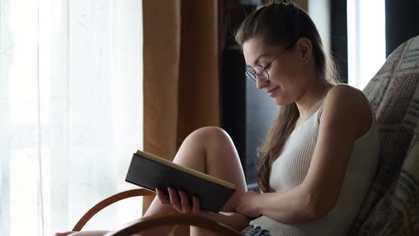 The girl in glasses reads a book in natural light near the window and chuckles