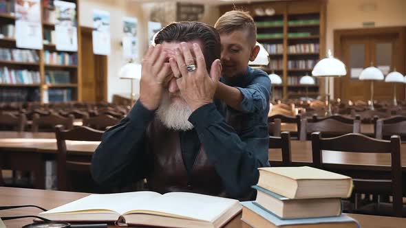 Teen Boy Closing Grandfather's Eyes with His Hands while He Reading Book in Library