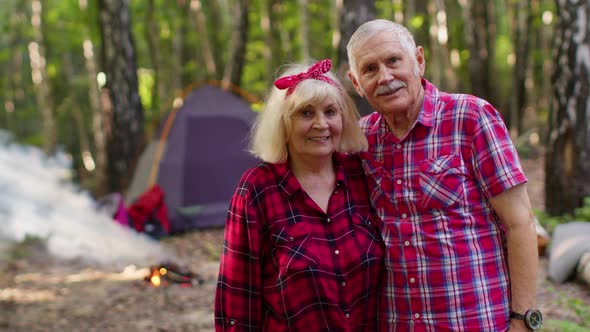 Portrait of Old Happy Couple Resting at Camping in Forest Over Campfire and Tent Background