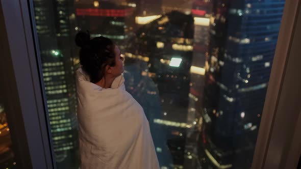 Attractive Woman Wrapped in a Blanket Looks Out the Window at the Lights of the Night City in a