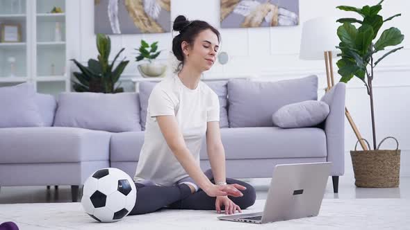 Young Woman Sitting in Front of the Laptop and Doing Relaxing Yoga Exercises on the Floor