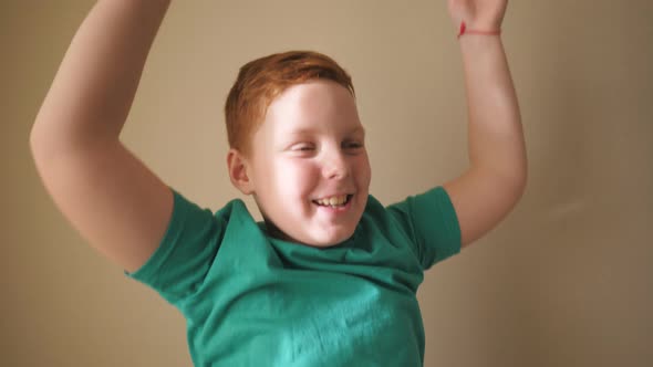 Happy Male Ginger Child with Freckles Looking Into Camera and Raises Hands Rejoicing Achievement