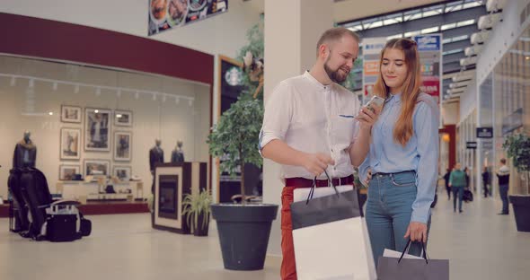 Happy Young Couple with Shopping Bags and Smartphone Talking in Mall