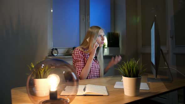 Shocked Female at Computer Talking on Smartphone. Sitting in Dark Room at Wooden Desk with Notebook