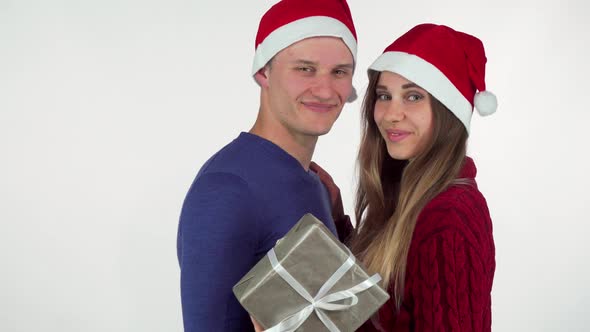 Young Loving Couple Wearing Christmas Hats Smiling To the Camera, Holding a Gift