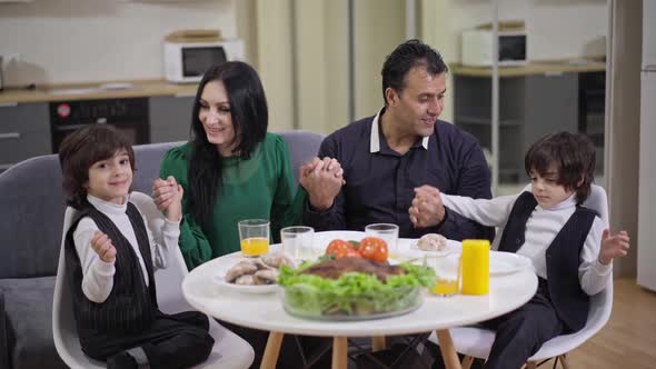 Happy Family Holding Hands Sitting Around Thanksgiving Dinner Table in Living Room at Home