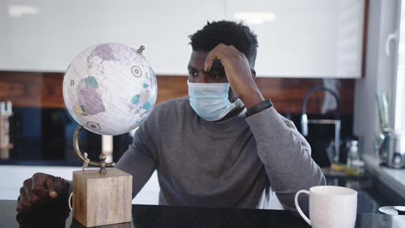 Lonely African American Black Man Traveler Looking at Globe in His Home with Medical Mask. Traveling