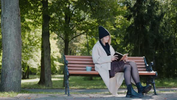 Young Girl Sitting on a Park Bench Reading a Book and Smiling in Autumn