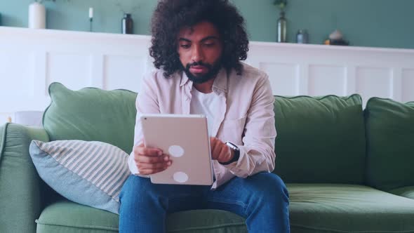 Young Curly Arabian Man Playing Game Through Electronic Tablet Sits on Sofa