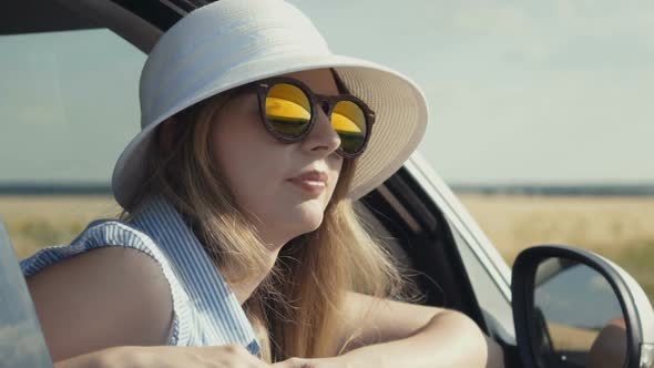 Happy Young Woman with Sunglasses and Hat Sitting in Car Passenger Seat and Looking Out Window on