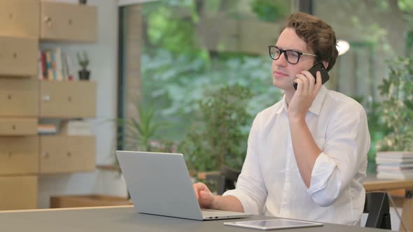 Young Man Talking on Smartphone While Using Laptop in Modern Office