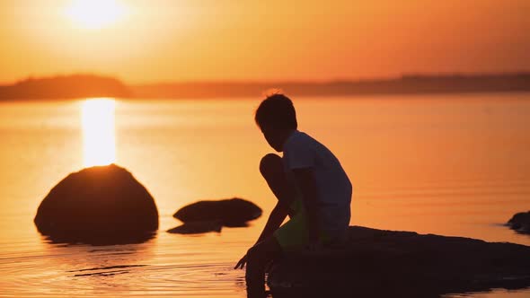 Little boy sitting on a stone among the water. Silhouette of a child near the river