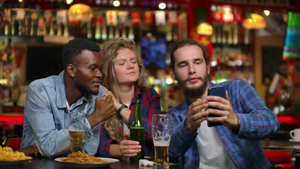 Two Men and a Woman Look at the Phone Screen While Sitting in a Bar and Prepare to Take a Shared