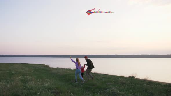 Happy Family Running with Flying Kite on Field Near Seashore During Sunset Slow Motion