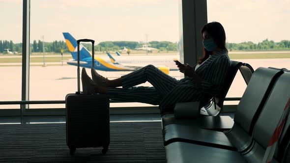 Woman in Mask, with Luggage, Uses Mobile, Sitting Against Panoramic Window with Runway and Big Plane