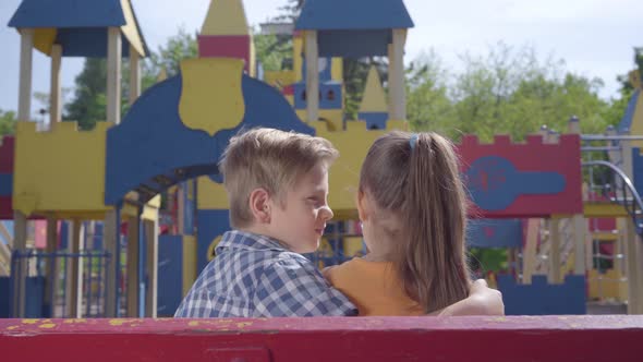 Cute Blond Boy Kissing Cheek of a Pretty Girl Sitting on the Bench in Front of the Playground. A