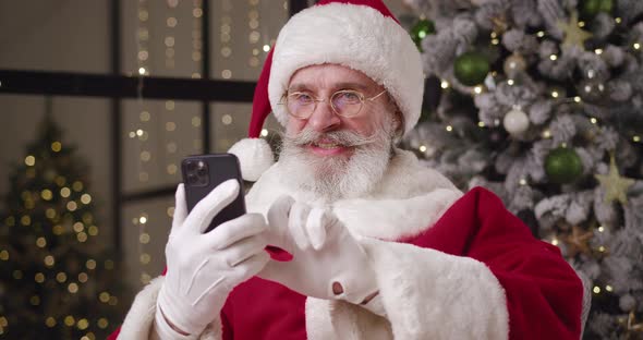 Closeup of Santa Claus Sitting Against Christmas Tree in His Residence Using Mobile Phone Swiping