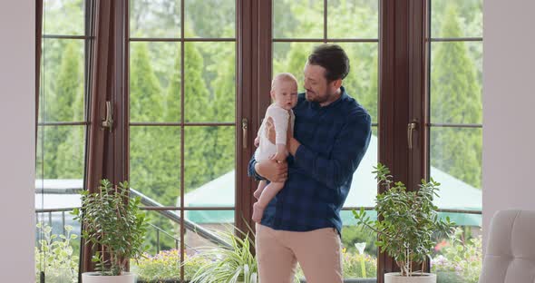 Bearded and Mustashed Father Holds His Adorable Baby and Slightly Waves on the Legs Smiling in Front