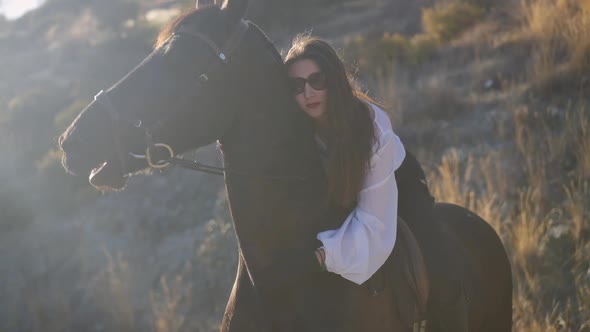 Brunette Caucasian Woman in Sunglasses Hugging Neck of Brown Horse Sitting on Back of Graceful