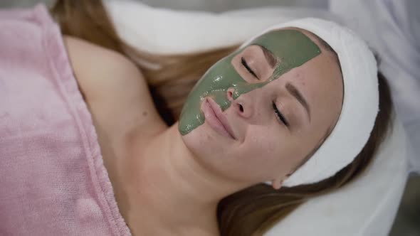 Healthy Young Girl with Green Clay on Face Opens Eyes and Relaxes with Smile