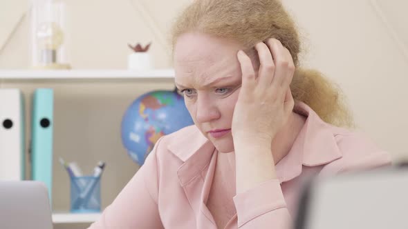 Close-up Face of Desperate Caucasian Senior Woman Holding Head with Hand As Looking at Laptop Screen