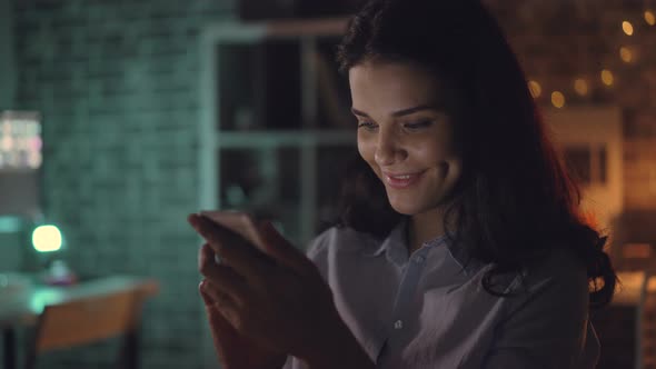 Cheerful Young Woman Using Smartphone at Night in Dark Office Smiling